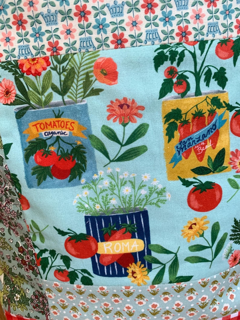 May include: A light blue fabric featuring a repeating pattern of potted tomato plants in various containers labelled Tomatoes Organic, San Marzano with Basil, and Roma, interspersed with orange and pink flowers and green foliage.
