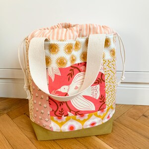 Op de afbeelding: Een tas met trekkoordsluiting en een patchworkontwerp met diverse patronen, waaronder zonnen, een witte vogel op een roze achtergrond, bloemenprints en een ruitjesmotief. De tas heeft cr&egrave;mekleurige stoffen hengsels en een gestreepte sluiting.