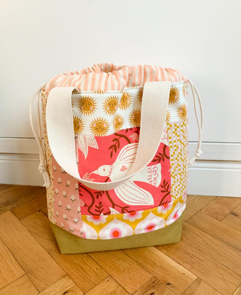Op de afbeelding: Een tas met trekkoordsluiting en een patchworkontwerp met diverse patronen, waaronder zonnen, een witte vogel op een roze achtergrond, bloemenprints en een ruitjesmotief. De tas heeft cr&egrave;mekleurige stoffen hengsels en een gestreepte sluiting.