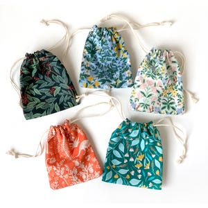 以下が含まれることがあります： 花柄の巾着袋が5つ。葉、ベリー、花など、緑、青、オレンジ、白の色合いで様々なデザインが施されています。各バッグには白い巾着の留め具が付いています。