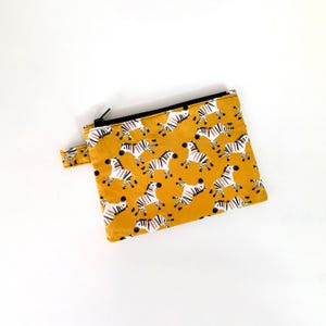 Puede incluir: Un estuche con cremallera de color amarillo mostaza con un estampado repetido de cebras a rayas negras, blancas y naranjas. El estuche tiene una cremallera negra y una pequeña presilla de tela en el lateral.