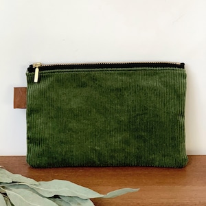 Può includere: Un astuccio rettangolare in velluto a coste verde foresta con cerniera dorata e una piccola linguetta in pelle marrone. L'astuccio è appoggiato su una superficie di legno. Il velluto a coste ha delle scanalature verticali.