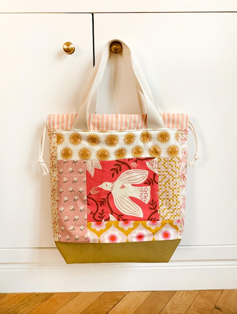 Op de afbeelding: Een patchwork draagtas met diverse stofpatronen, waaronder zonnemotieven, een witte vogel op een rode achtergrond, bloemenprints en roze strepen. De tas heeft twee cr&egrave;mekleurige hengsels, een trekkoordsluiting en een effen goudkleurige onderkant.