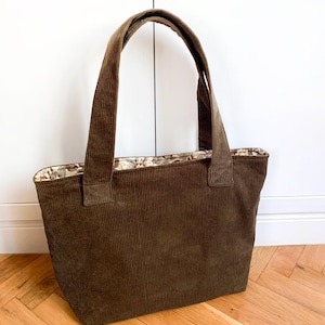 Op de afbeelding: Een donkerbruine corduroy tote bag met twee bijpassende schouderbanden. De binnenvoering is gemaakt van een lichte stof met een gedetailleerd bloemenpatroon in bruin-, blauw- en crèmetinten, zichtbaar langs de bovenrand van de tas.