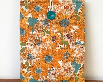 May include: A floral print book sleeve featuring a vibrant pattern of orange and yellow sunflowers with teal accents. The sleeve is secured with a teal button and a white cord loop. The background is white.
