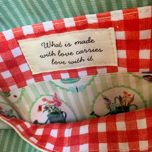 May include: A fabric pouch featuring an orange and white gingham border and a cream-coloured label with the text, What is made with love carries love with it. The interior lining displays a light green striped pattern with floral illustrations.