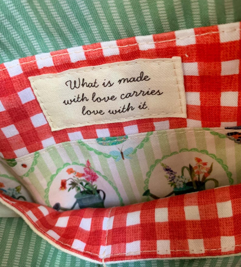 May include: A fabric pouch featuring an orange and white gingham border and a cream-coloured label with the text, What is made with love carries love with it. The interior lining displays a light green striped pattern with floral illustrations.