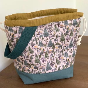 May include: A quilted drawstring bag featuring a light pink fabric with a repeating pattern of trees and small houses. The bag has a solid teal base, a mustard yellow top collar, a dark green fabric strap, and white cord drawstrings on both sides.