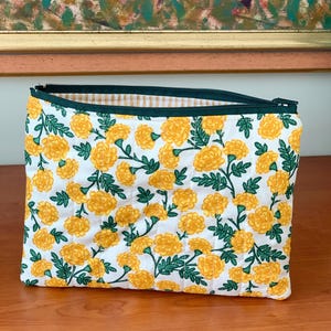 Może przedstawiać: Prostokątna, pikowana kosmetyczka z białym tłem i wzorem żółtych kwiatów nagietka i zielonych liści. Zamek błyskawiczny jest ciemnozielony, a podszewka wewnątrz ma wzór w żółto-białą kratkę.