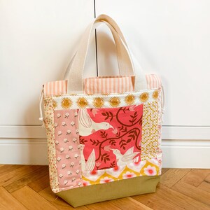Op de afbeelding: Een patchwork draagtas met een mix van stoffen, waaronder een roze bloemenprint, een rood paneel met witte vogels en ranken, een geel-wit geometrisch patroon en een effen olijfgroene basis. De tas heeft cr&egrave;mekleurige hengsels en een trekkoordsluiting.