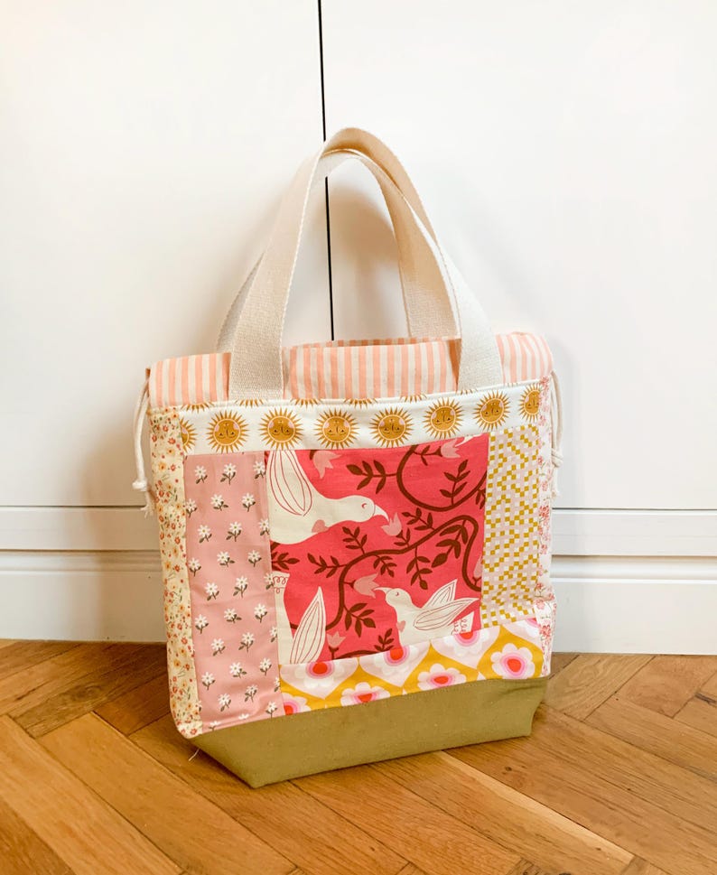 Op de afbeelding: Een patchwork draagtas met een mix van stoffen, waaronder een roze bloemenprint, een rood paneel met witte vogels en ranken, een geel-wit geometrisch patroon en een effen olijfgroene basis. De tas heeft cr&egrave;mekleurige hengsels en een trekkoordsluiting.