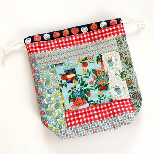 May include: A fabric drawstring bag featuring a patchwork design with various patterns including red strawberries on navy blue, red and white gingham, light blue floral prints, and a central panel depicting tomato plants and garden cans.