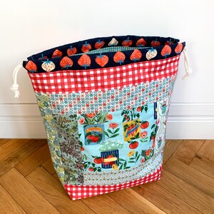 May include: A fabric drawstring bag featuring a patchwork design with red and white gingham, floral patterns, and a light blue panel printed with tomato seed packets and garden vegetables. The interior lining has a dark blue strawberry print.