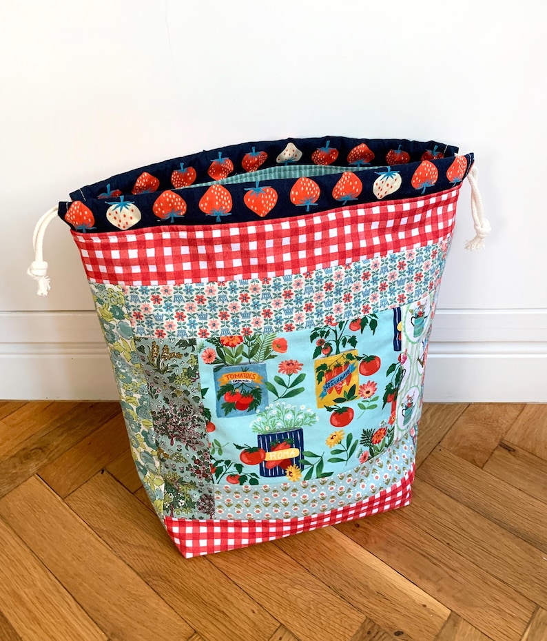 May include: A fabric drawstring bag featuring a patchwork design with red and white gingham, floral patterns, and a light blue panel printed with tomato seed packets and garden vegetables. The interior lining has a dark blue strawberry print.