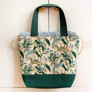 May include: A tote bag featuring a cream-colored fabric with a repeating pattern of peacocks, green foliage, and small blue flowers. The bag has dark green fabric handles, a solid dark green base, and a blue and white striped drawstring top closure.