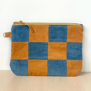 May include: A rectangular corduroy pouch with a checkerboard pattern in shades of blue and orange. The pouch has a gold zip and a small loop on the side. The corduroy fabric has vertical ridges.