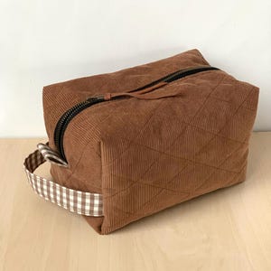 Op de afbeelding: Een bruine corduroy reistas met een gewatteerd patroon. De tas heeft een zwarte rits, een bruin leren ritstrekker en een handvat van bruin-wit geruite stof. De rechthoekige tas staat op een licht houten oppervlak.