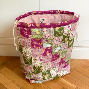 Puede incluir: Una bolsa acolchada con cordón ajustable que presenta un diseño de patchwork con patrones florales y botánicos en tonos magenta, verde oliva, rosa y blanco. La bolsa tiene un borde superior magenta sólido, cordones blancos y forro interior rosa.