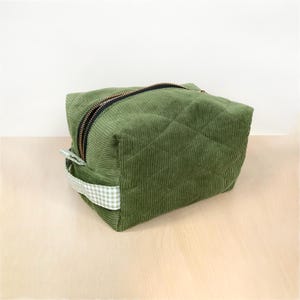 Op de afbeelding: Een groene corduroy make-uptas met een gewatteerd patroon. De tas heeft een zwarte ritssluiting en een klein handvat met een wit en groen ruitpatroon. De kubusvormige tas staat op een lichtgekleurd oppervlak.