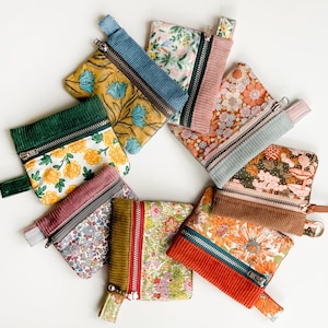 May include: A collection of small, zippered pouches in various floral patterns and corduroy accents. The pouches feature a range of colors, including yellow, blue, pink, and orange. Each pouch has a metal zipper and a small fabric loop.