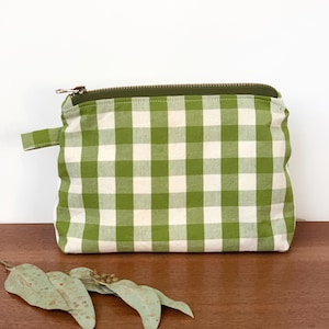Könnte beinhalten: Eine grün-weiß karierte Reißverschlusstasche. Die Tasche hat einen grünen Reißverschluss und eine kleine Stoffschlaufe an der Seite. Die Tasche liegt auf einer Holzoberfläche.