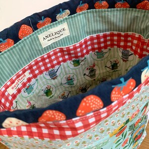 May include: A fabric drawstring bag featuring a navy blue exterior with a red strawberry pattern. The interior is lined with a red and white gingham fabric and a light green striped fabric. A label reads ANELIQUE Made to Carry Joy.