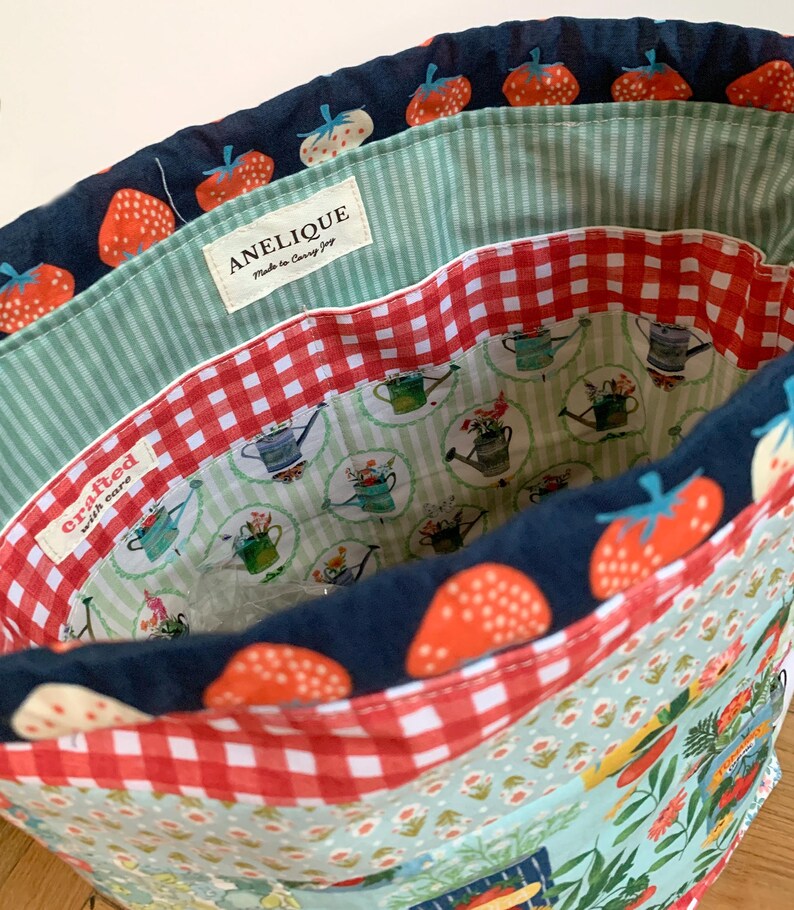 May include: A fabric drawstring bag featuring a navy blue exterior with a red strawberry pattern. The interior is lined with a red and white gingham fabric and a light green striped fabric. A label reads ANELIQUE Made to Carry Joy.