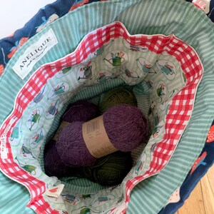 May include: A fabric drawstring bag with a dark blue exterior featuring a strawberry print. The interior is lined with light green striped fabric, a red and white gingham border, and a pattern of watering cans. Inside are several balls of yarn.