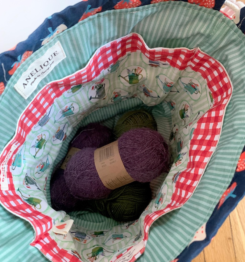 May include: A fabric drawstring bag with a dark blue exterior featuring a strawberry print. The interior is lined with light green striped fabric, a red and white gingham border, and a pattern of watering cans. Inside are several balls of yarn.