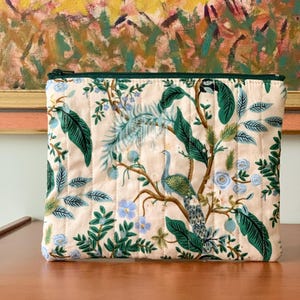 May include: A rectangular quilted pouch with a cream background and a peacock and floral pattern. The design features green, blue, and cream colours. The pouch has a green zip and is on a wooden surface.