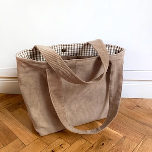 Op de afbeelding: Een beige corduroy tote bag met twee bijpassende schouderbanden. De binnenzijde is gevoerd met een bruin-witte gingham ruitjesstof en is voorzien van een enkele metalen drukknoop in het midden om de opening van de tas te sluiten.