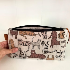Op de afbeelding: Een rechthoekig beige etui met ritssluiting met een herhaald patroon van bruine en zwarte kattenillustraties. De etui heeft een zwarte ritssluiting en een bruine leren treklip. De binnenkant is lichtbeige.