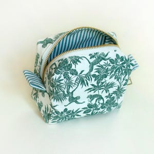 Può includere: Un beauty case bianco con stampa giungla verde con scimmie e fogliame. La borsa ha una cerniera dorata e un interno a righe blu e bianche. La borsa è aperta.