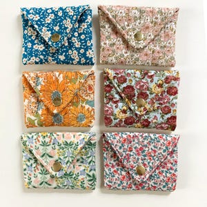 May include: Six small, square fabric wallets with floral patterns. Each wallet has a flap closure secured by a gold-colored snap. The patterns include blue and white, pink, orange, red, and green floral designs. The wallets are arranged on a white surface.