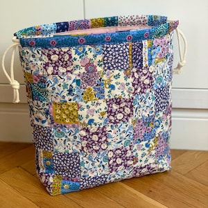 Puede incluir: Una bolsa acolchada con cordón que presenta un diseño de patchwork con varios patrones florales en tonos morado, azul, amarillo y blanco. La bolsa tiene un forro interior rosa claro y cordones de algodón blanco grueso a ambos lados.