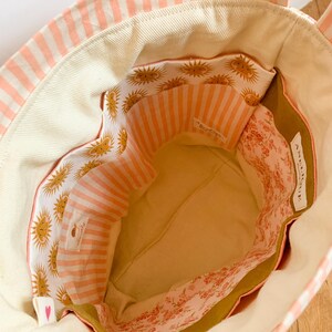 Op de afbeelding: Een stoffen draagtas met een cr&egrave;mekleurige binnenkant, een zak met zonnepatroon en een zak met roze bloemenprint. De tas heeft een perzik en wit gestreepte buitenkant en een wit label met de tekst ANGELIQUE.