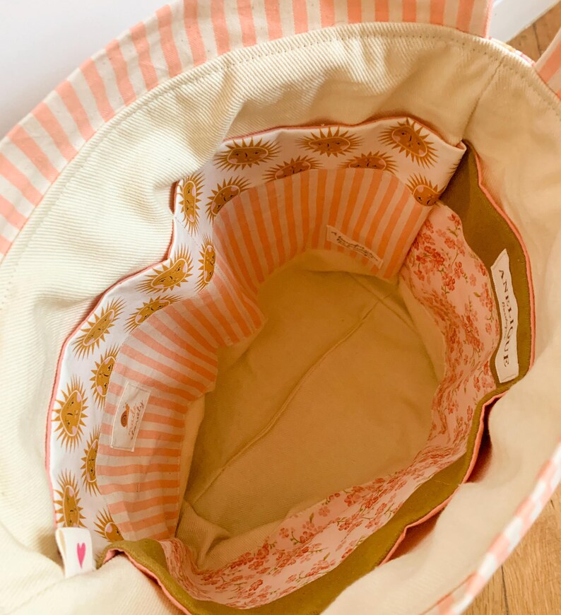 Op de afbeelding: Een stoffen draagtas met een cr&egrave;mekleurige binnenkant, een zak met zonnepatroon en een zak met roze bloemenprint. De tas heeft een perzik en wit gestreepte buitenkant en een wit label met de tekst ANGELIQUE.