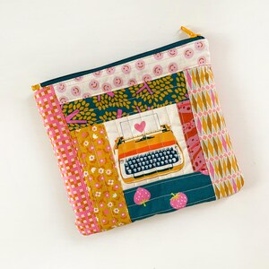 May include: A rectangular quilted zipper pouch featuring a patchwork design with various fabric patterns including pink smiley faces, teal leaves, yellow flowers, strawberries, and a central graphic of an orange typewriter with a pink heart.