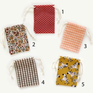 May include: Five rectangular fabric drawstring pouches labeled one through five. Pouch one is red with white hearts. Pouch two has a floral pattern. Pouch three is peach gingham. Pouch four is brown gingham. Pouch five is mustard yellow with zebras.
