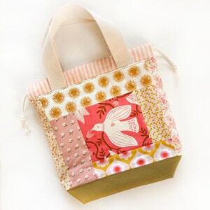 Op de afbeelding: Een patchwork tas met trekkoord en een centraal paneel met een witte vogel op een roze achtergrond, omringd door secties met bloemen-, zon- en geometrische patronen in roze, geel en olijfgroen, met cr&egrave;mekleurige canvas handvatten.