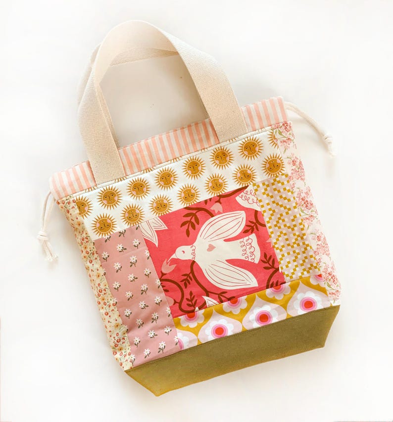 Op de afbeelding: Een patchwork tas met trekkoord en een centraal paneel met een witte vogel op een roze achtergrond, omringd door secties met bloemen-, zon- en geometrische patronen in roze, geel en olijfgroen, met cr&egrave;mekleurige canvas handvatten.