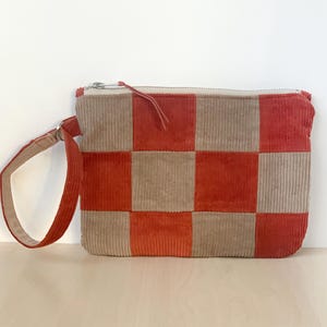May include: A corduroy clutch bag with a red and tan checkerboard design. It features a zip closure and a matching red handle. The rectangular bag has a soft, textured appearance.