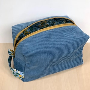 May include: A blue corduroy wash bag with a floral patterned lining. It features a yellow zip and a small handle with a floral design. The rectangular bag is on a light wooden surface. The bag is ideal for storing toiletries.
