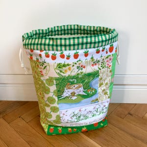 May include: A fabric drawstring bag featuring a patchwork design with various nature-themed patterns, including strawberries, green foliage, and a central illustration of a frog sitting on a tree stump. The top edge has a green and white gingham trim.