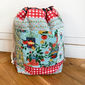 May include: A fabric drawstring bag featuring a patchwork design with red and white gingham, light blue floral patterns, and a central print of tomato plants in cans with the text La Rosa. The bag has cream-colored cotton rope drawstrings.