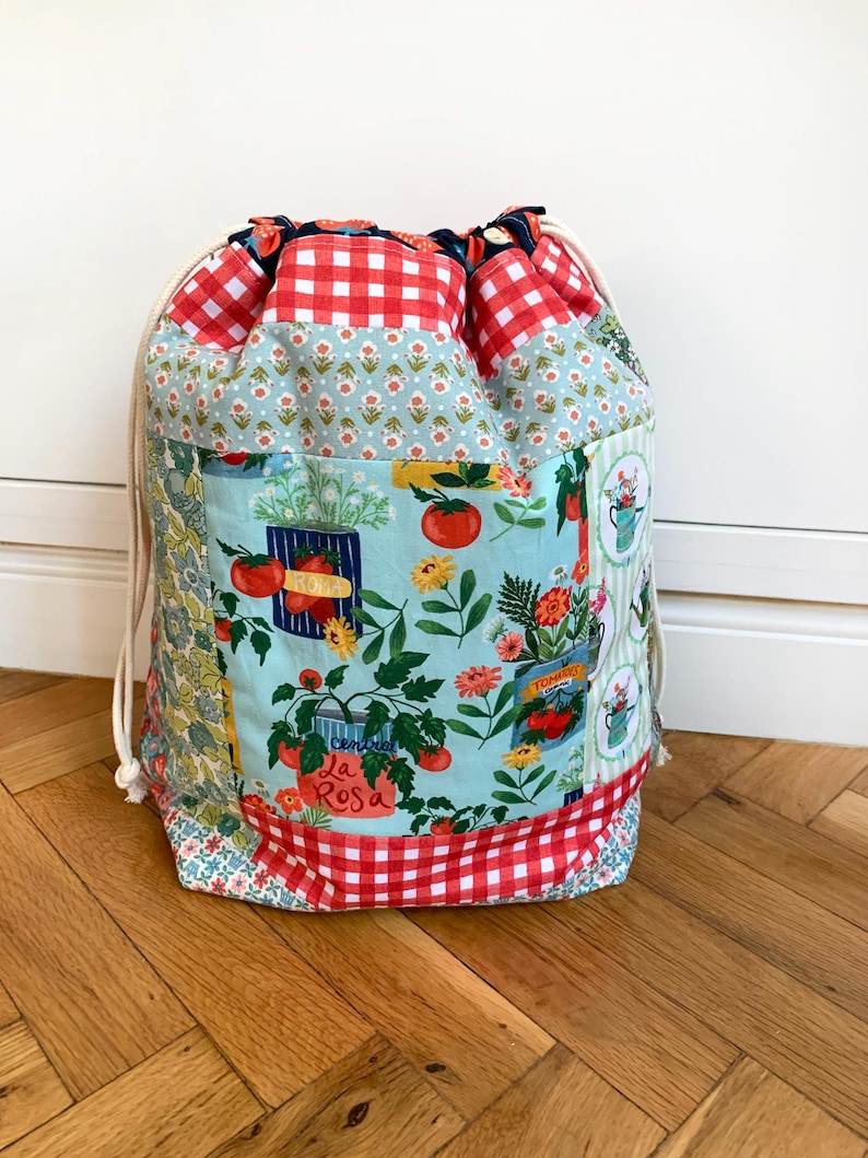 May include: A fabric drawstring bag featuring a patchwork design with red and white gingham, light blue floral patterns, and a central print of tomato plants in cans with the text La Rosa. The bag has cream-coloured cotton rope drawstrings.