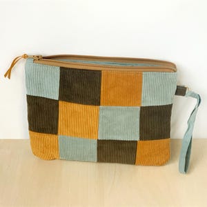 May include: A corduroy patchwork clutch in a square pattern of brown, mustard yellow, and teal green. The zippered pouch has a brown zipper and a teal wrist strap. The bag is sitting on a light wood surface.