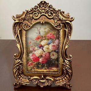 Könnte beinhalten: Vergoldeter Bilderrahmen mit floralem Gemälde. Der Rahmen hat aufwendige Verzierungen und ein detailliertes Design. Das Gemälde zeigt einen Strauß roter, rosa, weißer und blauer Blumen. Der Rahmen steht auf einer braunen Oberfläche.