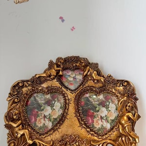 May include: Ornate gold-coloured picture frame with three heart-shaped apertures, each showing a floral print. The frame features cherub figures and floral embellishments. The text "Grandma Frame" is visible in the upper left corner.