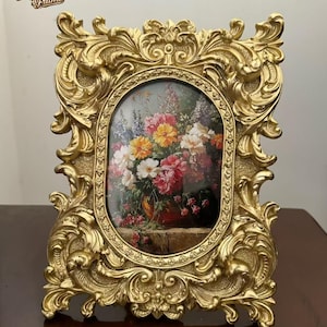 May include: An antique-style gold picture frame with an oval aperture, displaying a floral painting. The frame has an elaborate, baroque design with swirling patterns. The text "Grandma Frame" is visible in the upper left corner.